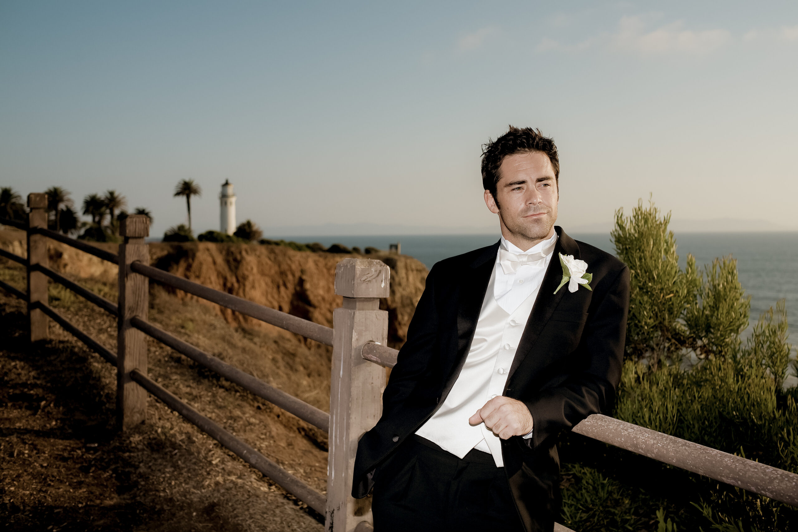 groom leaning on a fence by the ocean with Palos Verdes Lighthouse in the background during a California wedding