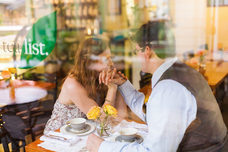 Couple seen through window at The Tea List in Davis, intimate candid engagement moment with reflections and soft light